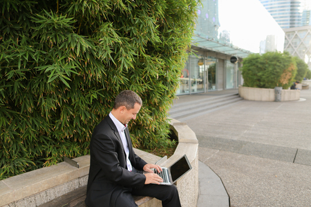 Businessman typing by laptop outside near green plant in   and looking at watch. Concept of ecology thinking for business. Successful man dressed in black suit working with computer outdoors.の写真素材