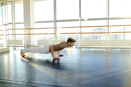 Interpreter doing press exercises in hall, young man fond of sports since childhood. Fair-haired tattooed guy in white trousers lie on parquet floor near ballet barre. Concept of sportswear, well-equipped gym for workouts or gymnastic school.の写真素材