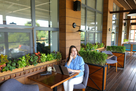 Female student chatting with friends by laptop at cafe. Young girl dressed in jeans shirt sitting near window with room plants. Concept of communicating with close people and modern technologies.の写真素材