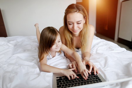 sweet portrait of family using laptop laying on white bed woman and kid girl daughter with sunshine. concept of new technologies or happy parent, motherhood and maternityの写真素材