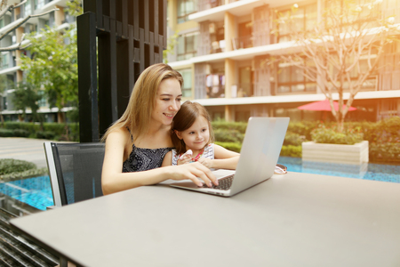 happy parent mother and daughter using laptop on background of swimming pool during sunny day. concept of social networks new technologies, family time recreation joy or relationship, advertising of english courcesの写真素材
