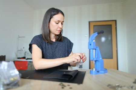 Female slicing leather using special knife, ruler and scissors, young lady recently got job in atelier. Craftswoman with neat red manicure standing near wooden table with tools in well-lit workshop and preparing materials for making shoes. Concept of accuracy, handicraft or fine work.の写真素材