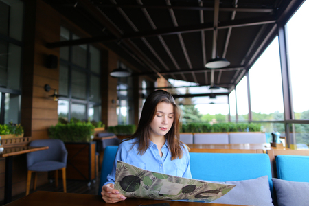 Gorgeous female person reading menu on sofa at cafe with black notebook on table  . Young woman wears blue shirt and has cute smile. Concept of cheap prices restaurants.の写真素材