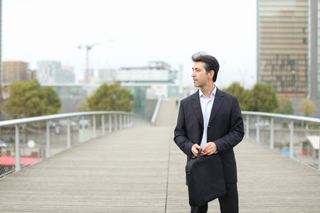 Lawyer in business clothes with smartphone waiting for client, man standing on bridge checking time on silver gadget. Middle-aged American with bag wearing strict suit looking by sides. Concept of fashionable man outfits, law services or innovative technologies.の写真素材