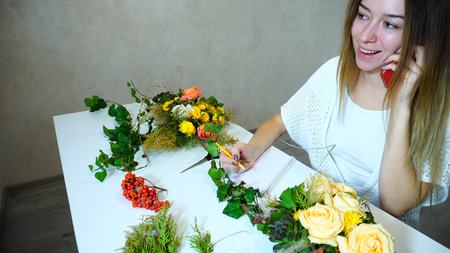 Sweet girl  talking on cell phone with client and clarifies order of bouquet of roses, records information about meeting in notebook and talks pleasantly, gives recommendations for care of flowers, sitting at table in bright office in daytime. Girl with long blond hair  dressed in white sweater. Concept of profession and favorite work, gadgets and technology, communication on phone with customers, floristry and design of flowers in form of beautiful compositions of plants, successful girl landscapers.の写真素材