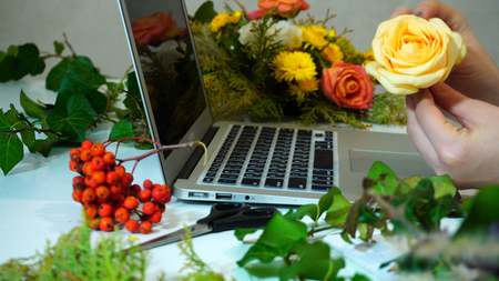Shooting close-up of hands of marvelous female florist who holds delicate bud of pale yellow beautiful rose in hands, puts it on table and type text on computer keyboardの写真素材