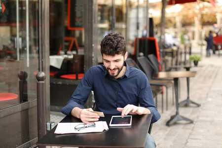 Gladden prosperous man purchasing at online shop with money card and tablet with close up face in  . Satisfied man sitting at street cafe and smiling. Concept of buying goods using Internet.の写真素材