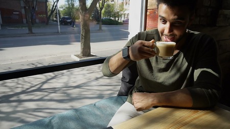 Handsome man sits near window and drinks coffee. Guy has short hair, beard and wears smart watch on hand. Boy dressed in khaki pullover. Concept of modern technologies for businessmen caf for business meetings and tasty coffee.の写真素材