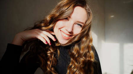 Female Smiling, Looking at Camera, Laughs or Flirts. Girl With Long Curly Brown Hair Sitting in White Room With Christmas Lights and New Year Chloe on Background. Scene Filmed by Sun at Day.の写真素材