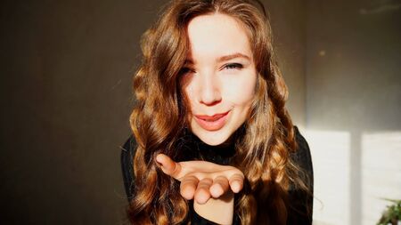 Female air kiss Smiling, Looking at Camera, Laughs or Flirts. Girl With Long Curly Brown Hair Sitting in White Room With Christmas Lights and New Year Chloe on Background. Scene Filmed by Sun at Day.の写真素材
