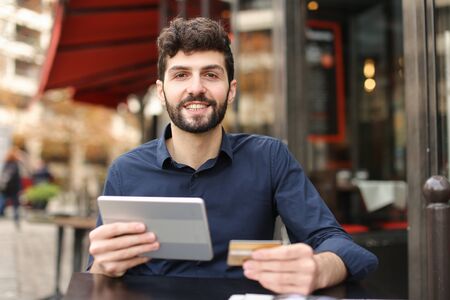 Traveler purchasing tickets and paying with money card by tablet in  . Happy man sitting and cafe and wears classic style blue shirt. Concept of cheap tours and online booking.の写真素材