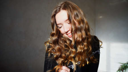 Female Smiling, Looking at Camera, Laughs or Flirts. Girl With Long Curly Brown Hair Sitting in White Room With Christmas Lights and New Year Chloe on Background. Scene Filmed by Sun at Day.の写真素材