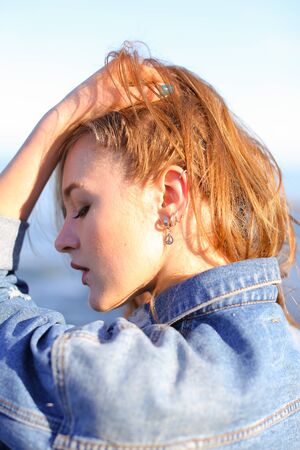 Attractive young woman with smile on face turns head to sides and develops beautiful hair, turns in profile and poses, holding hands on belt and stands with back to camera lens on sea shore against blue sky on clear summer day.の写真素材