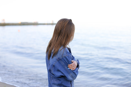 Attractive young woman walks slowly and walks along sea shoreline and examines neighborhood of beach with smile on face. European-looking girl with medium-length blond hair dressed in black pants, white T-shirt with bright pictures on top of which blue denim jacket and on sea beach on warm evening.の写真素材
