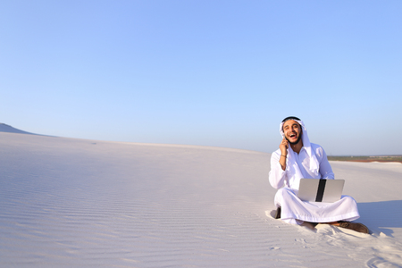 Modern male Arab and builder dials on cellular customer to clarify stages of construction and sits with computer on white clean sand outdoors on hot summer day.の写真素材