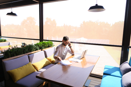 Successful sales manager checking result diagrams, working with laptop and calling marketer indoors. Male person typing number on smartphone looks assured and confident in white shirt. Concept of selling goods at online store and setting consumers.の写真素材