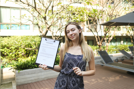 woman is holding pen and contract, proposes to sign document against background of building with apartments. concept of signing papers, travel rental housing. Front used with Open Font Licenseの写真素材