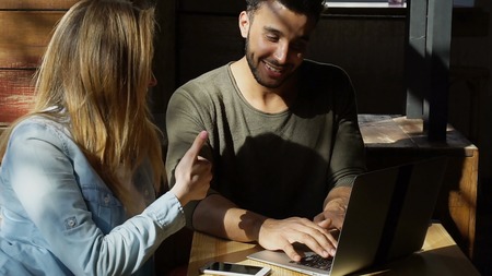 On table modern gray laptop and white smartphone. Bearded man with dimples shows something woman in...