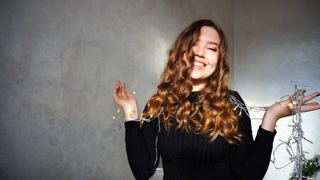 Female Smiling, Looking at Camera, Laughs or Flirts. Girl With Long Curly Brown Hair Sitting in White Room With Christmas Lights and New Year Chloe on Background. Scene Filmed by Sun at Day.の写真素材