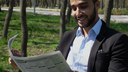 Arabian guy sits on chair near table and reading newspaper. Man dressed in blue shirt, white jeans, black jacket and brown shoes. Concept of news in newspapers interesting facts about celebrities and correct facts.の写真素材