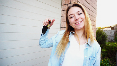 Cheerful woman standing near garage with car keys. Concept of purchasing car and happy owner. Young female person wears blue jeans shirt.の写真素材