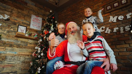 Fervent little boys enthusiastically holding holiday boxes. Three beautiful guys give gifts to camera, rejoice and smile. Toddlers  guess what surprise Santa has prepared.  children of European-looking brothers dressed in warm sweaters  blue jeans with socks, sit on large  soft armchair  cozy room decorated  Christmas decorations, fashionable floor lamp, walls  which New Years posters hang,  high  tree, under  lie   Surprises   tied  ribbons. Concept  traditions  family happy    Year, laughter  carefree childhood  friendly siblings, interior desired     positive emotions  eve    holidays, active pastime  children foolishness,  attributes  symbols.の写真素材