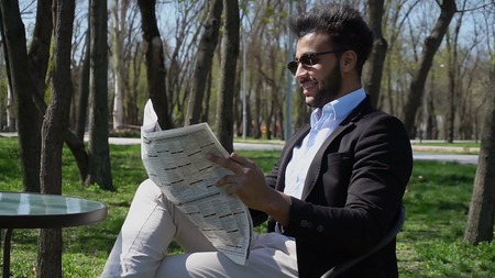 Guy sits on chair near table and reading newspaper. Man has beard, short hair and dimples. Male dressed in black jacket, white jeans and blue shirt. Concept of newspapers advertisement places for reading quite clean parks day off.の写真素材