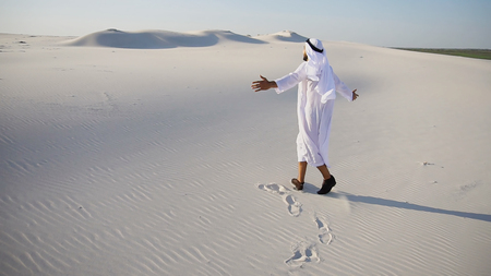 Cheerful Muslim guy Arabian UAE Sheikh walks along white sand of wide desert on warm summer evening. Arab young man in national clothes dances on spot and laughs giggly, adjusting hands with harness in wind. Swarthy, handsome Muslim with short dark hair dressed in kandura, long, spacious dress made of white unpainted cotton with knitted lace cap of hafia, on top of which tied goutrail and dark brown shoes. Concept of Arab and Muslim man, united Arab emirates and beautiful landscapes, sheikh in desert and solitude with nature, Emirati national clothes, good mood and happy emotions.の写真素材
