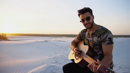 Romantic bearded young Arab man exercises musical instrument with strings, smiles and becomes comfortable in position to start playing guitar, standing in middle of wide sandy desert on warm summer evening at sunset. Swarthy man with dark hair and short haircut in sunglasses dressed in camouflage t-shirt, black pants and brown shoes. Concept of handsome Arab young man, hobby or favorite thing to do, learning or playing stringed musical instruments, good mood and positive emotions, beautiful scenery and amazing travel.の写真素材