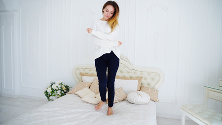 Cute Female European Appearance Jumps at Full Length on Soft and Big Bed and Fooling Around and Smiling Relieves Stress After Hard Day and Wearing White Sweater and Dark Blue Pants is Bright and Spacious Bedroom. Concept of Beautiful and Successful Young People, Promotion of Womens Beauty or Facilities Stylish Youth Clothes, Smiles and Emotions, Luxury Apartments and Decor.の写真素材