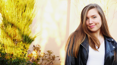Pretty smiling girl passing with close up face near house. Concept of spring weather and happy female person. Young woman wears black leather jacket and white shirt.の写真素材