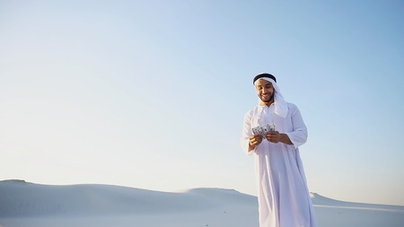 Stately young male Arab man in good mood conducts financial training of money, smiles and laughs joyfully launches money to wind, standing in middle of bottomless desert with white sand on sunny summer day. Swarthy Muslim with short dark hair dressed in kandura, long, spacious dress made of white, unpainted cotton with knitted lace cap, gafia, over which headscarf tied. Concept of Arab and Muslim men, business and finance, smile of fortune and great mood, united Arab emirates and beautiful landscapes, national clothes of Emirates.の写真素材