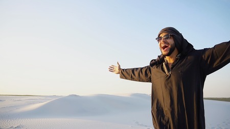 Attractive young sheikh with smile on face looks into camera and goes forward, stops and takes off glasses, enjoys and shows expanses of bottomless desert with white clean sand and clear blue sky on summer evening. Swarthy Muslim with short dark hair and sunglasses dressed in black kandur, long spacious dress made of dark cotton with knitted lace cap, gafia over which headscarf and dark brown shoes are tied. Concept of Arab and Muslim men, united Arab emirates and beautiful landscapes, advertising services of travel companies or airlines, national clothes of Emirates, good mood and enjoyment of life.の写真素材