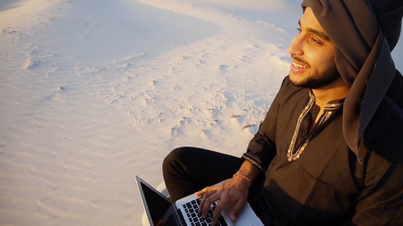 Joyous male Arab sits on sand at computer. young man engaged in scientific work or writes article typing on keyboard of gadget, relaxes sitting in silence of bottomless desert against blue sky on summer evening. Swarthy Muslim with short dark hair and beard dressed in black kandur, long spacious dress made of dark cotton with knitted lace cap of hafia, on top of which headscarf and dark brown shoes are tied. Concept of Arab and Muslim men, education and modern technology, business and entrepreneurial activities, Emirati national clothing, good mood and enjoyment of life.の写真素材
