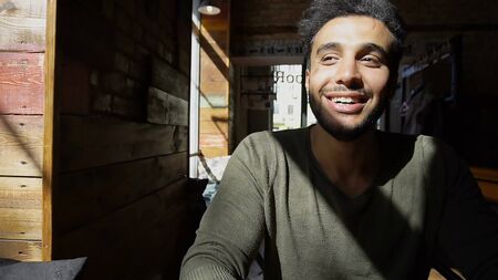 Muslim guy sits in caf and looking at window, put hand on table. Happy boy has dimples, beard, full lips and black hair. Person dressed in khaki pullover and wears watch on hand. Concept of modern places good atmosphere cheap coffee.の写真素材
