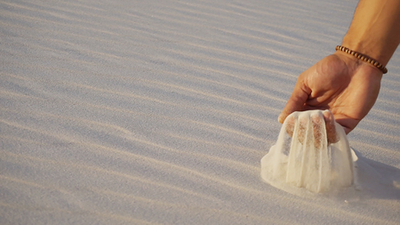 Close-up of face and hands of young Muslim man Arabian Sheikh who takes sand into hand and blows small white grains of sand through fingers, guy smiles and looks out into distance of bottomless desert on hot summer day. Swarthy, handsome Muslim with short dark hair dressed in kandura, long, spacious dress made of white unpainted cotton with knitted lace cap of hafia, on top of which tied goutrail and dark brown shoes. Concept of Arab and Muslim men, united Arab emirates and beautiful landscapes, peace and tranquility, national clothing of emirates.の写真素材
