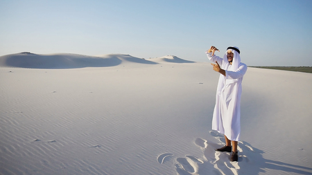 Successful young Arabian UAE Sheikh businessman male Muslim uses laptop to build drawing in Photoshop and prints fingers on laptop sitting on white sand in bottomless wide desert in afternoon against blue sky. Swarthy Muslim with short dark hair dressed in kandura, long, spacious dress made of white unpainted cotton with knitted lace cap of hafia, on top of which headscarf and dark brown shoes are tied. Concept of Arab and Muslim men, business and business, modern technology and gadgets, united Arab emirates and beautiful landscapes, sheikh in desert and seclusion from nature, national clothes of emirates.の写真素材
