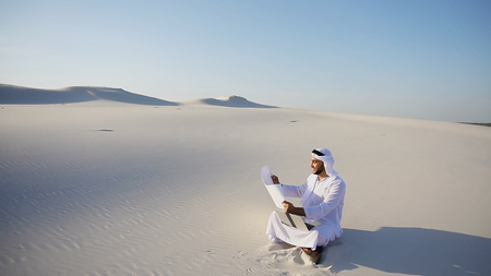 Handsome Arabian UAE Sheikh Arab businessman and construction worker uses computer for design, busy developing complex schemes and opens drawing for checking and clarifying details sitting on white sand in bottomless wide desert in afternoon against blue sky. Swarthy Muslim with short dark hair dressed in kandura, long, spacious dress made of white unpainted cotton with knitted lace cap of hafia, on top of which headscarf and dark brown shoes are tied. Concept of Arab and Muslim men, business and business, modern technology and gadgets, united Arab emirates and beautiful landscapes, sheikh in desert and seclusion from nature, national clothes of emirates.の写真素材