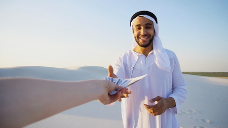 Portrait of emirate male advertising agent or bank employee who, with smile on face, attracts and advertises goods or services of bank, gestures and concludes deal, receives bills that are handed by hand from camera, standing in middle of bottomless sandy desert with white clean sand against blue sky with hot In evening outdoors. swarthy, handsome Muslim with short dark hair dressed in kandura, long, spacious dress made of white unpainted cotton with knitted lace cap of hafia, on top of which tied goutrail and dark brown shoes. Concept of Arab and Muslim men, United Arab Emirates and beautiful landscapes, advertisement of travel company or airline advertising, national clothes of Emirates, good mood and happy emotions.の写真素材
