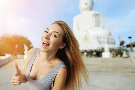 happy joyful girl showing like or thumbs up, background of Big Buddha mountain Phuket, traveling to sunny Thailand. Concept of movement of people in relatively large geographical locationsの写真素材