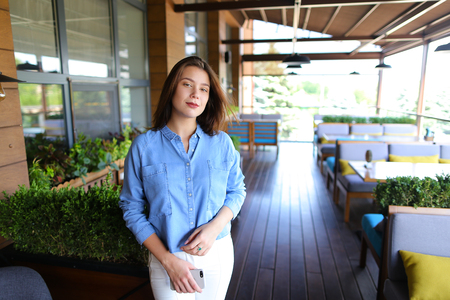 Businesswoman standing at cafe with smartphone and wearing jeans shirt and white pants. Concept of fashionable youth and modern technology.の写真素材