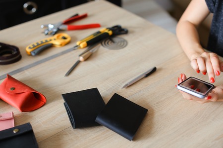 Female hands at leather atelier using smartphone, handmade wallets and tools on table. Concept of...