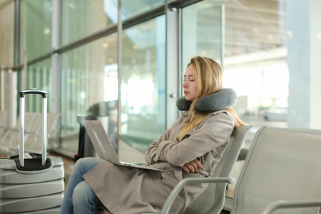 Pretty woman sitting in waiting room with laptop and using social networks near valise, using neck pillow. Concept of traveling and modern technology.の写真素材