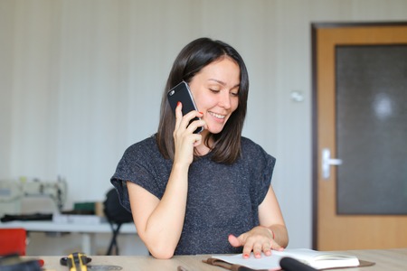 Beautiful woman speaking by smartphone and creating handmade leather notebook at home. Concept of handicraft, hobby and home business.の写真素材