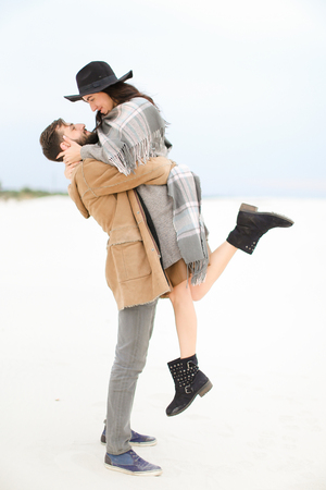 Man picking up young woman wearing hat, coat and grey scarf on white snow background. Concept of gladden couple and seasonal photo session.の写真素材