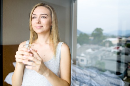 Young caucasian woman standing with glass of water at hotel in morning. Concept of healthy way of...