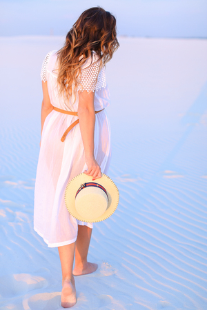 Back view of standing young woman on white blue sand and keeping hat. Concept of photo session in desert and summer vacations.の写真素材