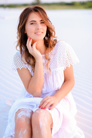 Young beautiful female person sitting on white sand and wearing dress. Concept of beauty and resting on beach, summer seasonal photo session.の写真素材
