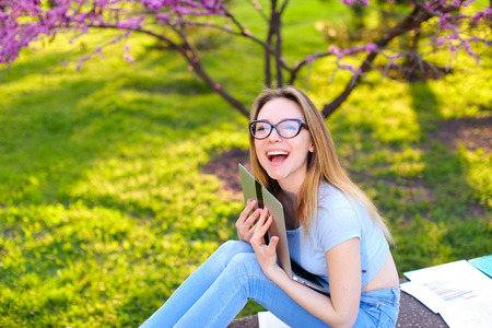 Smiling woman in glasses hugging laptop and sitting on grass in garden. Concept of modern gadgets...