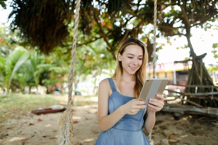 Young girl using tablet and riding swing on sand, wearing jeans sundress. Concept of summer vacations and modern technology.の写真素材